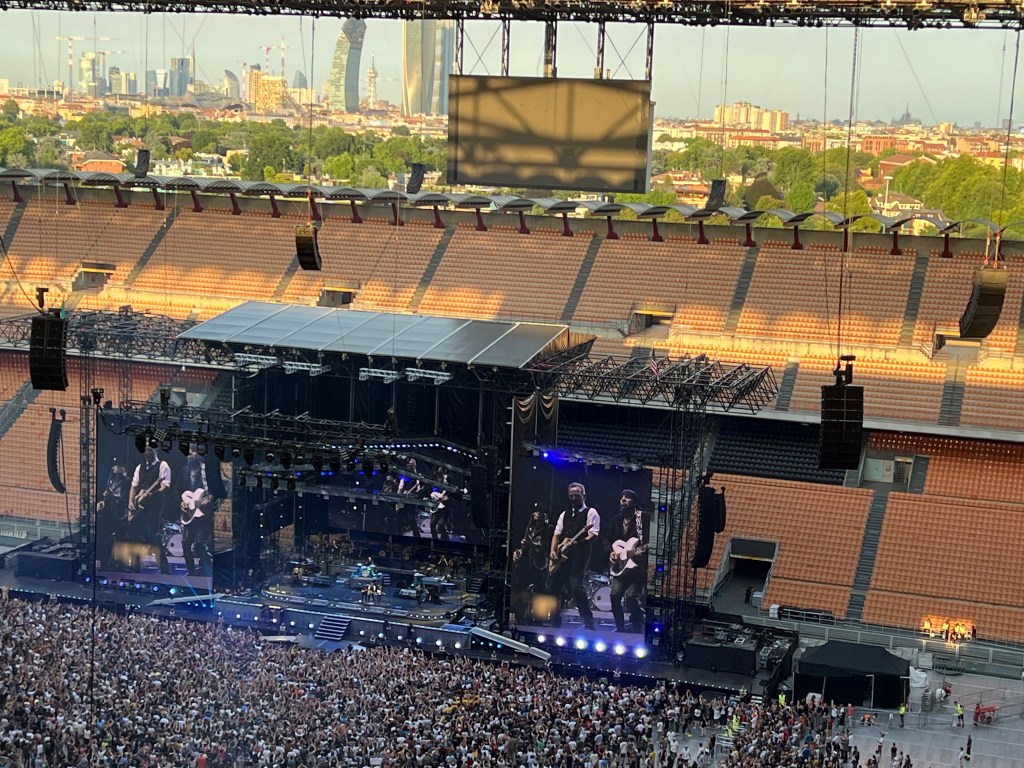 Concerto di Bruce Springsteen allo stadio San Siro, Milano, con vista sulla città sullo sfondo. La scena mostra il palco, i musicisti e una folla di fan entusiasti.